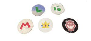 Collection of 5 circular wood disks with tactile features, each depicting a character logo from the Mario game. The badges show Mario's red M, Luigi's green L, Yoshi's egg, Princess Peach's crown, and Bowser's face.